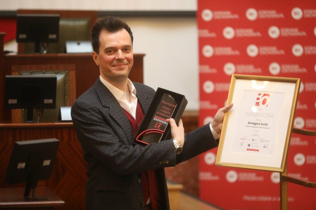 Chop we siwym szakecie, wiśniowym cwitrze i biołym hymdzie pokazuje do fotografije stauetka i dyplōm.